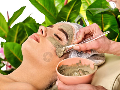 水疗沙龙中女人的泥面膜在治疗室用粘土全脸按摩去除皱纹在绿色植物背景下应用具有碗治疗背景图片