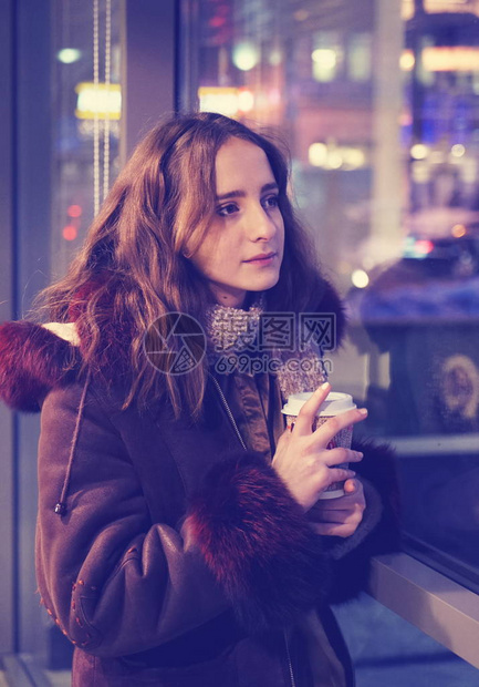 一个穿着带兜帽的保暖夹克的年轻女孩用纸杯和热咖啡暖手城市生活模图片