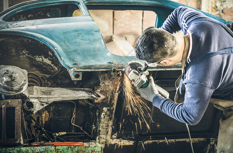 年轻人机械工人在凌乱的车库里用电动格栅修理旧的老式汽车身带有防护磨损的工作安全与使用技术设备的人一起使用的利基图片