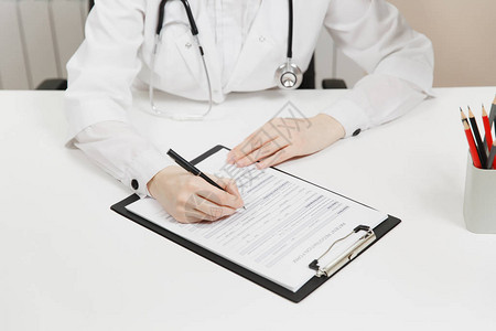 在医院的轻型办公室里工作填写医疗文件的医生手的裁剪镜头女人在咨询室桌子上的剪贴板上写在纸上医学概念顶视背景图片