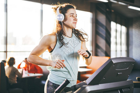 主题是运动和音乐一个漂亮的充气女人在跑步机上的健身房里跑步她头上戴着白色的大耳机背景图片