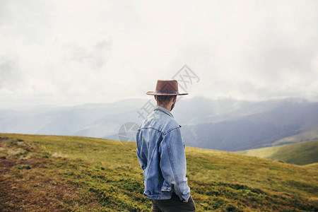 时尚旅行者戴着帽子站在阳光明媚的山上云层之上文本的空间在山上旅行的时髦家伙惊人的大气时刻旅行图片