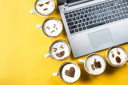 在黄色背景的笔记本电脑旁边的卡布奇诺杯子上涂着微笑的emoji情感和图片