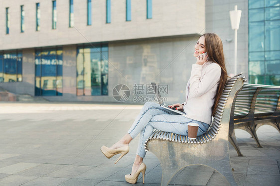 快乐的女商人在笔记本电脑上工作女保险代理人在电脑上查看电子邮件在手机上交谈坐在现代办公中心附近的户外长椅图片