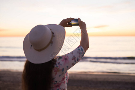 年轻妇女用手机或智能手机数码相机拍摄海和日落时的照片图片
