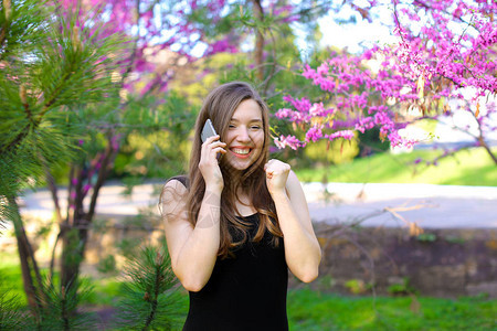 快乐的女人们通过智能手机与男朋友在开花果树附近的公园中交谈图片