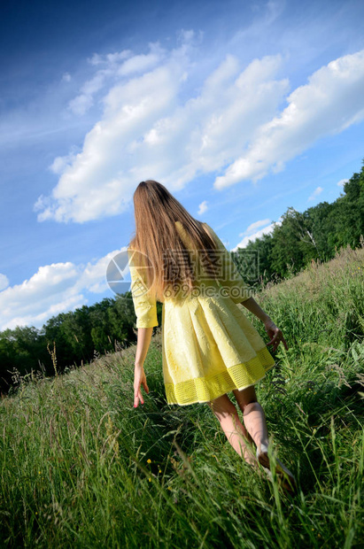 身穿黄色裙子的年轻女享受在草地上行走青草地有野花和蓝天图片