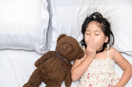 小女孩打哈欠睡在床上与泰迪熊娃保图片