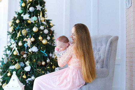 坐在椅子上带着孩子穿着粉红色礼服的年轻女子在圣诞树附近坐着图片