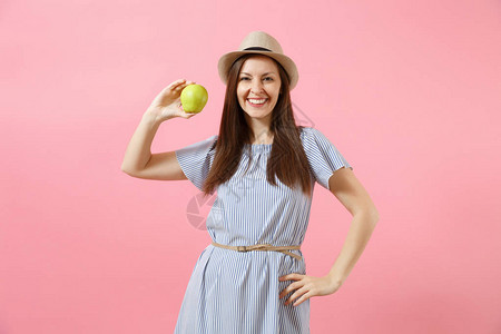 身穿蓝色连衣裙戴着夏季草帽吃着粉红色背景中突显的绿色新鲜苹果实的年轻漂亮女人的画像图片