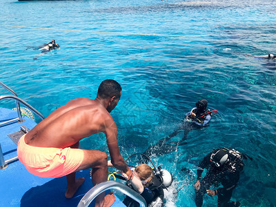 一个黑黝的阿拉伯运动健将帮助穿着黑色水肺潜水服的潜水员带着氧气瓶沉入大海中透明的蓝色海水下图片