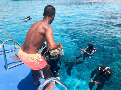一个黑黝的阿拉伯运动健将帮助穿着黑色水肺潜水服的潜水员带着氧气瓶沉入大海中透明的蓝色海水下图片