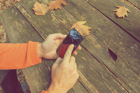 男手用智能手机在木制桌子上使用智能手机与秋天隔着户外图片