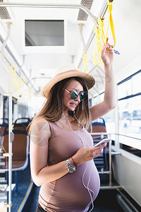 美丽的年轻孕妇站在城市公交车上看手机图片