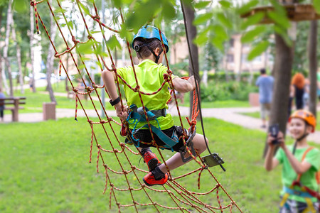 在冒险公园的绳索墙上攀爬安全设备的小男孩在智能手机上拍照的恶魔儿童夏季运动极限户外背景图片