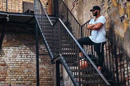 穿着运动服戴着帽子留着胡须的非裔美国人街球运动员拿着篮球站在楼梯上图片