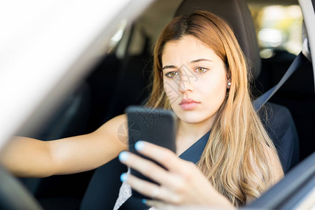 坐在汽车驾驶座上看着手机阅读短信的有吸引力的年轻女子的肖像注图片