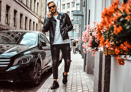戴着墨镜穿着黑色皮夹克和牛仔裤的时尚男士微笑着站在欧洲老街上的一辆豪华轿车旁图片
