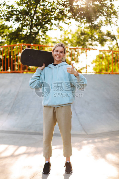 穿着连帽衫的漂亮笑脸女孩肩上拿着滑板快乐地看着镜头露出图片