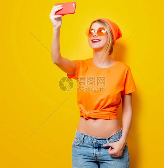 戴橙色眼镜的年轻女子用黄色背景的手机拍自图片