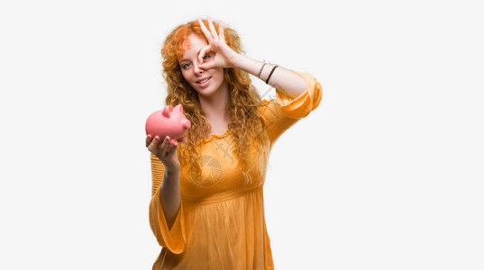 年轻红发女人拿着小猪银行带着快乐的笑脸微笑做一个好标记用手对着图片