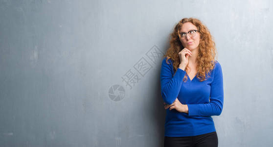年轻的红发女人在灰色的垃圾墙上用手托着下巴思考问题背景图片