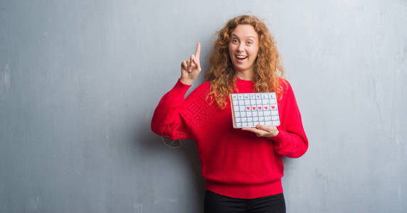 灰色格朗基风墙上的年轻红发女人拿着周期日历图片