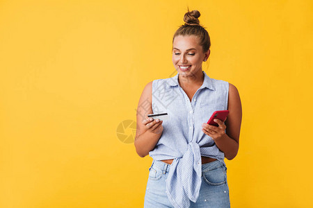 穿着衬衫的年轻快乐女人愉快地拿着信用卡和手机在黄色背景上手握图片