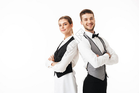 年轻的微笑服务员和穿着白衬衫和背心的漂亮女服务员背靠站着图片