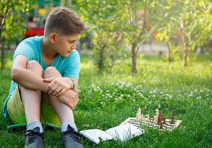 穿着绿色T恤的可爱男孩在坐草地上的公园棋盘上下象棋教育课图片