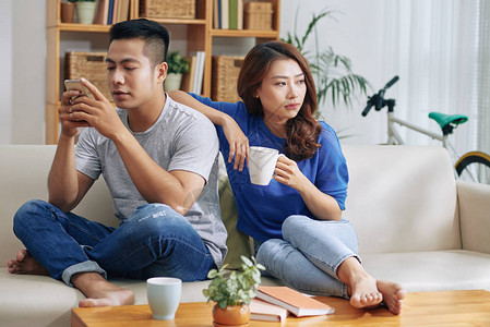 亚洲情侣坐在沙发上男人在智能手机上冲浪而背景图片