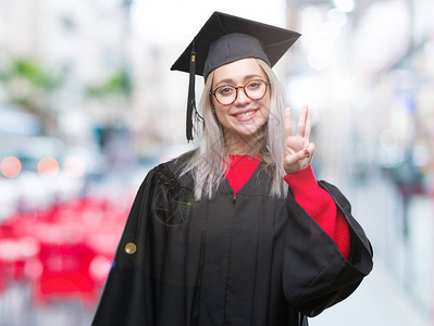 身穿毕业制服的金发年轻女郎在孤立的背景展出和指着三号手指时微笑着自信图片