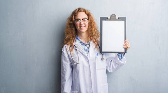年轻的红发女医生在灰色的格朗基墙上展示剪贴板图片
