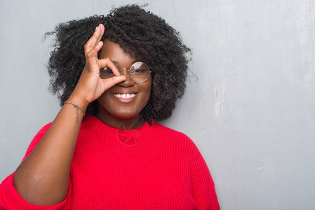 年轻的非洲裔美国妇女在灰色的垃圾墙上穿着冬季毛衣和眼镜做着很好的手势图片