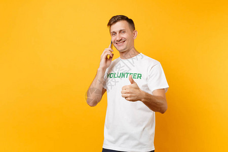 年轻人肖像在白色t恤与书面题词绿色标题志愿者谈手机孤立在黄色背景自愿无偿帮助背景图片