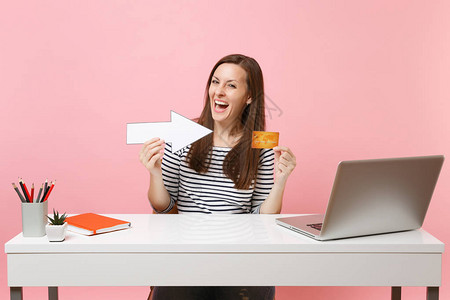 身穿散装衣用箭指着信用卡的欣喜妇女坐在办公室工作图片