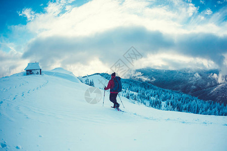 一个穿着雪鞋和登山杖的人在山上冬季旅行以云彩为背景的攀登山者游客找到了避难所山上的房子图片