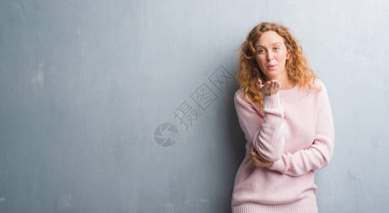 穿着粉红色毛衣的红发女青年在灰色长墙上站着看着摄影机用手亲吻空气图片