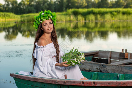 穿着白色复古连衣裙的黑色长发若虫坐在河中央的一艘船上头发上有一圈绿叶和白图片