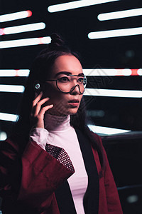穿着勃艮第和服无线耳机的迷人亚洲女孩在晚上霓虹灯下望向街道图片