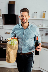 在厨房里拿着杂货袋和一瓶红酒的年轻男子长着英俊笑着图片