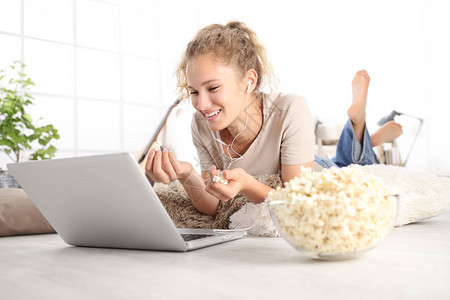 美丽的年轻微笑女人在上看电影吃爆米花躺在客厅的木地图片