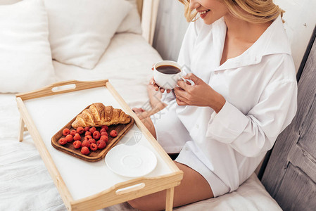 笑女孩坐在床上时拿着咖啡杯和餐盘早图片