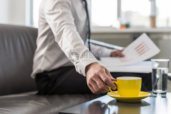 坐在办公室沙发上的商人伸手去拿他的黄色咖啡杯图片