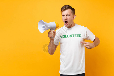 身穿白色T恤的男子肖像写着绿色志愿者在黄色背景上隔离的公共广播扩音器中尖叫自愿无偿帮助图片