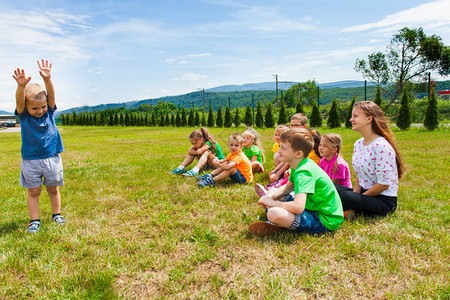 金发美女男孩站在一群坐在草地上的孩子面前举起手来图片