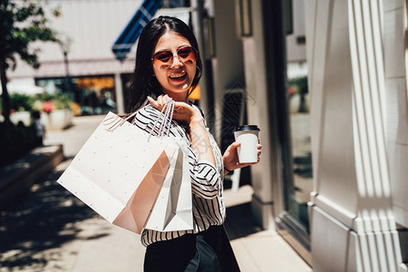 快乐的年轻亚洲女孩兴高采烈地面对镜头大笑戴着太阳眼镜的女人在阳光下拿着咖啡袋在中心商场出口户外购物站在街边图片