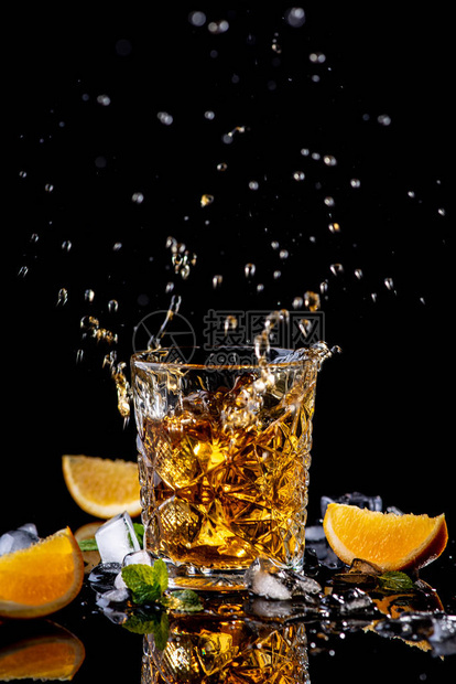 威士忌飞溅杯用橙色绿色薄荷和冰块装饰的威士忌在带有反射的黑色背景上图片