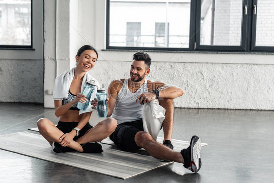 穿着服坐在瑜伽垫子上在健身锻炼后举瓶水的年轻夫妇微笑图片