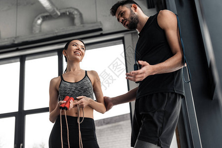 低角度的笑运动式夫妇与跳绳在健身房站立和说话图片